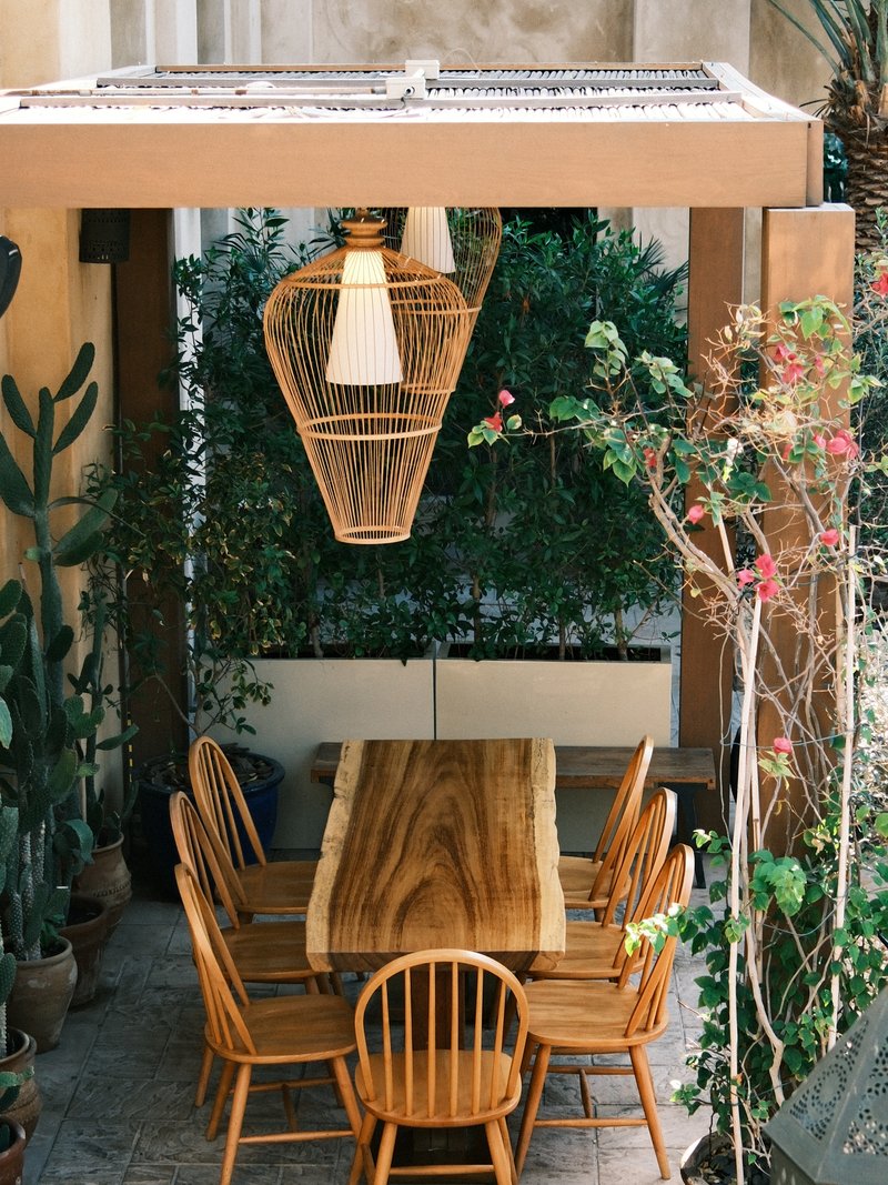 A wooden dining table surrounded by chairs under a pergola with hanging light and greenery.