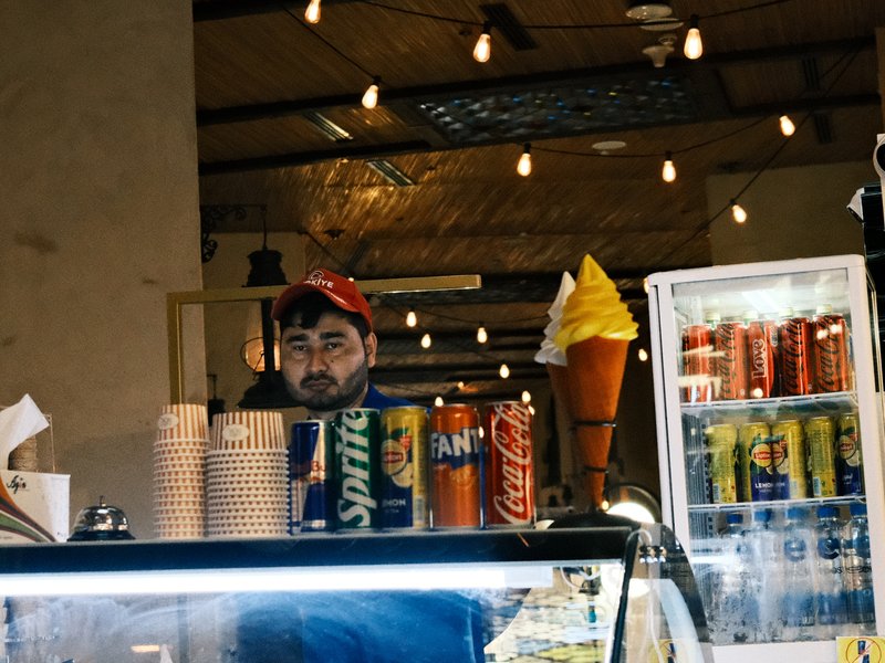 A man behind a counter with soft serve ice cream and various beverage cans in a cozy indoor setting.