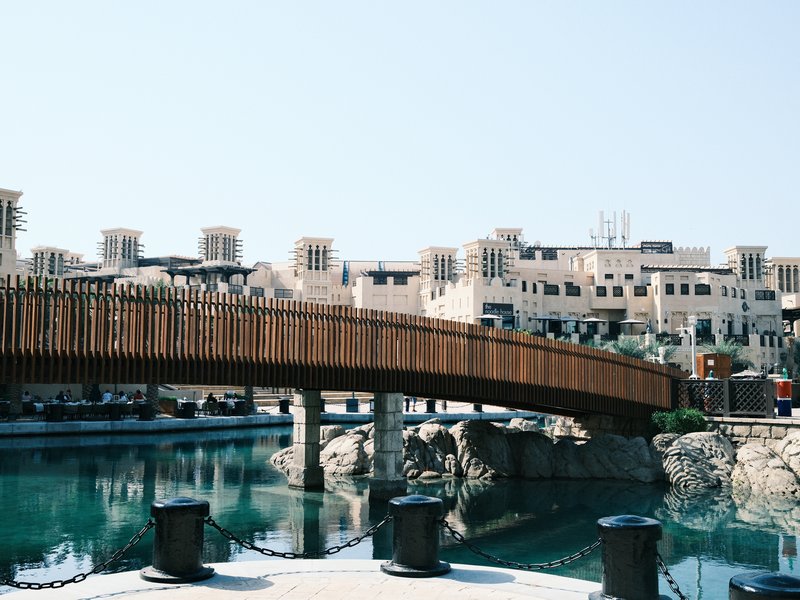 A wooden bridge over a calm water body with buildings in the background under a clear blue sky.