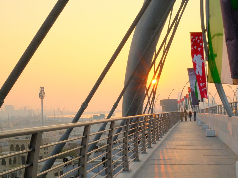 A sunset view from a modern pedestrian bridge with flags and silhouettes of people walking.