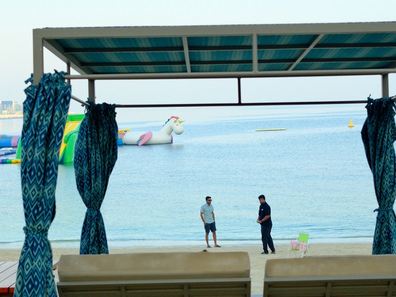 Two people standing on a beach under a shaded structure, with colorful inflatable structures in the water.