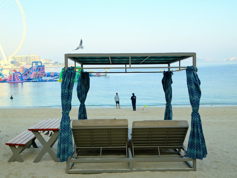 Two beach loungers under a canopy facing the ocean, with people walking along the shore in the background.