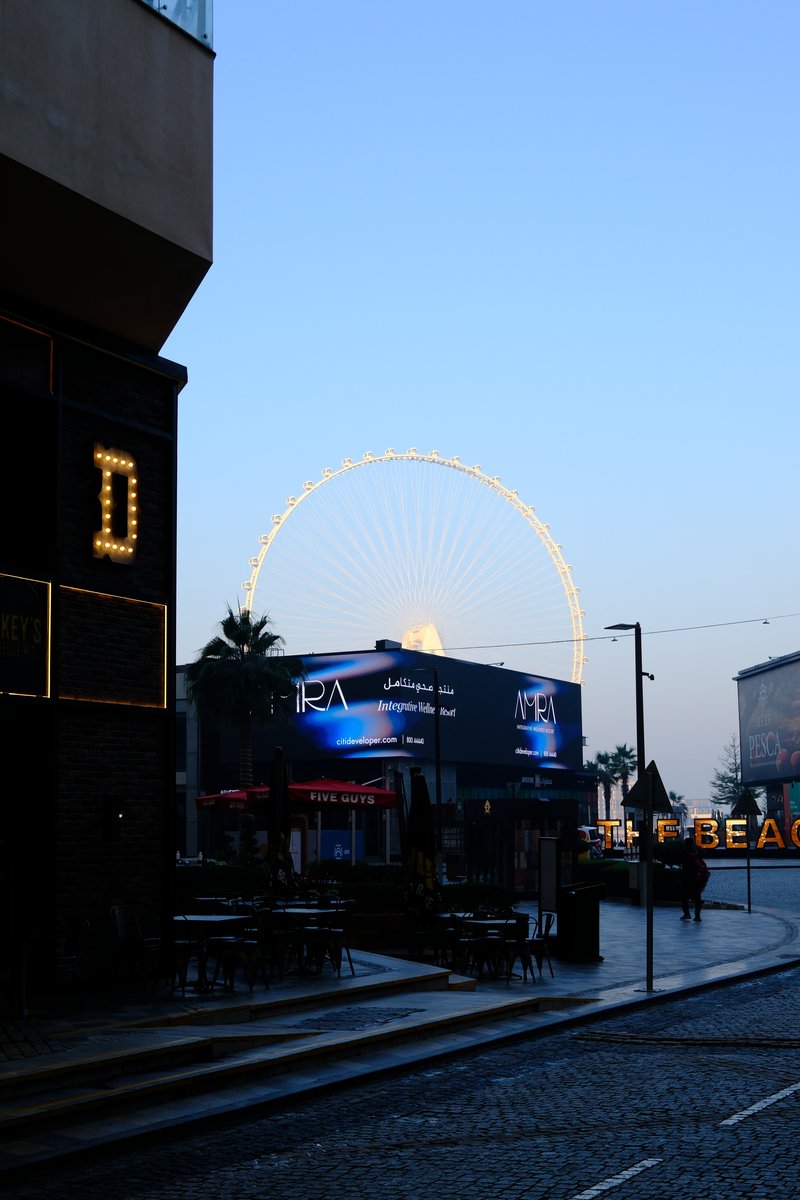 A street scene at dusk featuring a ferris wheel in the background and illuminated signage.
