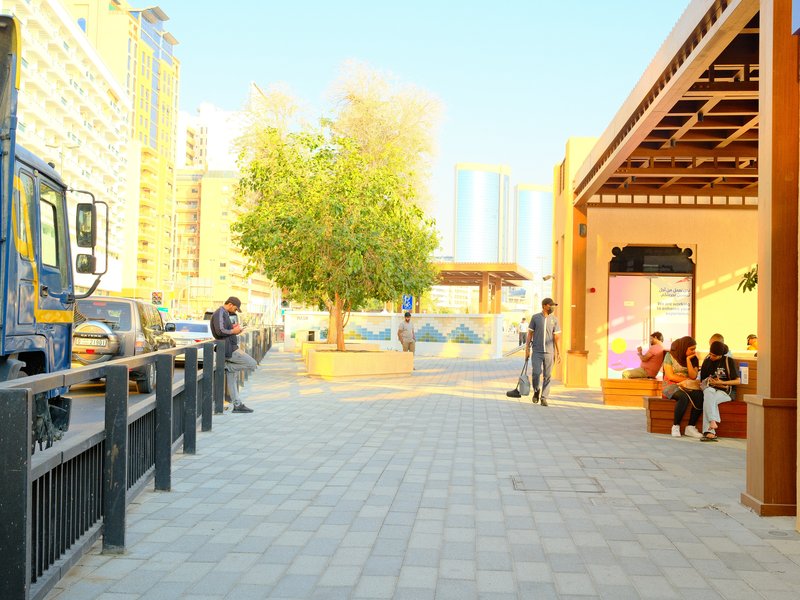 A vibrant urban scene with people interacting near a tree and modern buildings under bright sunlight.