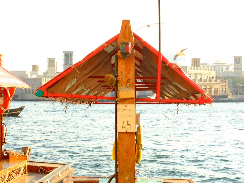 Wooden boat canopy with red roof over water, city skyline in background under bright sunlight.