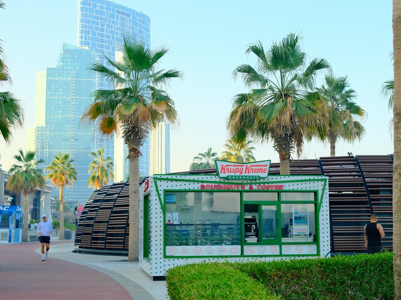 A green and white kiosk with palm trees and modern buildings in the background, set in a coastal urban area.