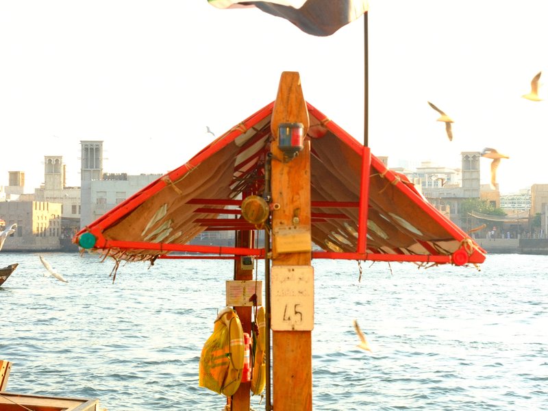 A colorful wooden structure by the water with birds flying around and a city skyline in the background.