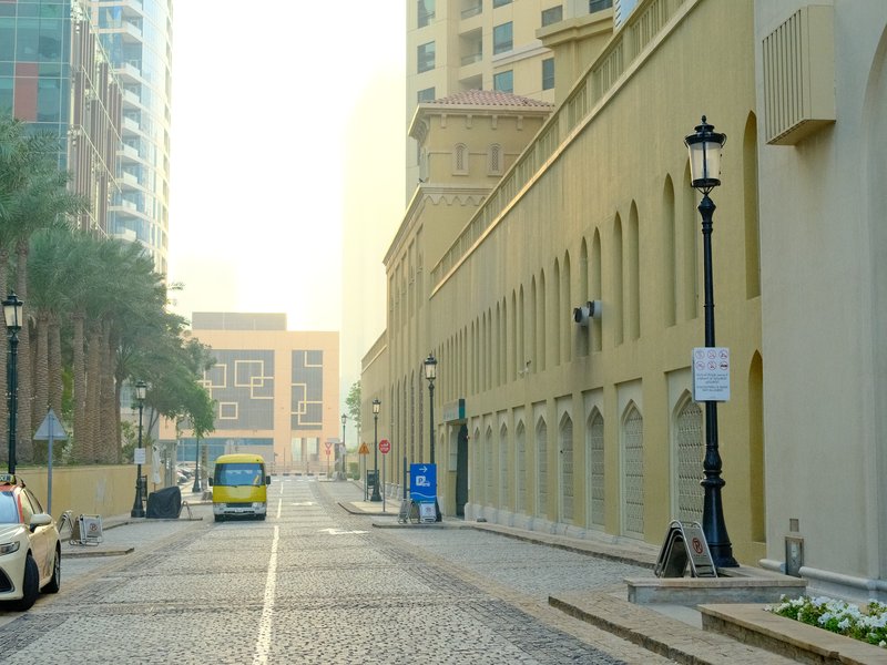 A quiet urban street with a light haze, featuring a cobblestone road, palm trees, and a yellow bus.