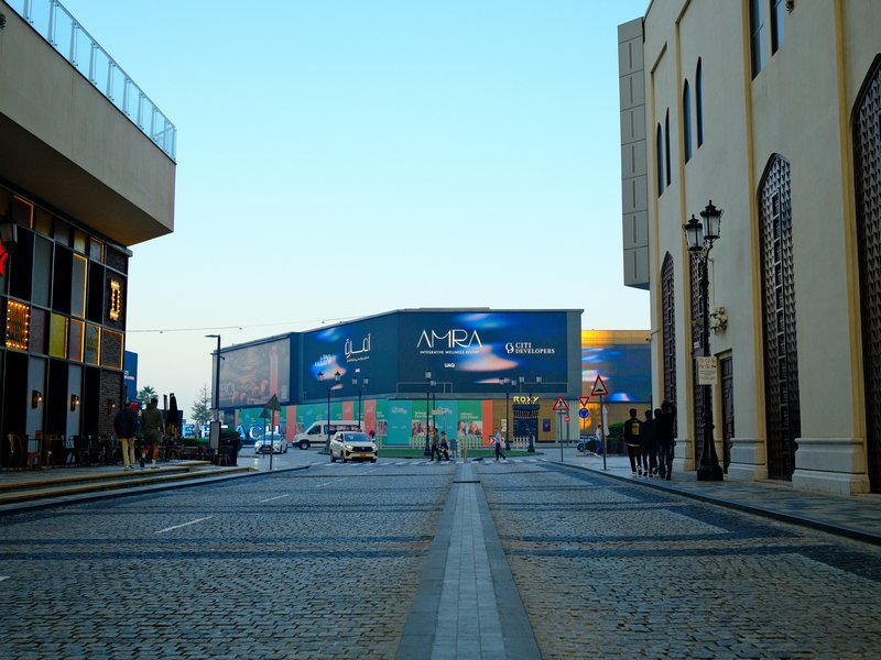 A street view with buildings on either side and a large billboard in the distance under a clear sky.