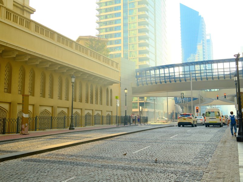Urban street scene with buildings, a pedestrian bridge, and vehicles under soft light.