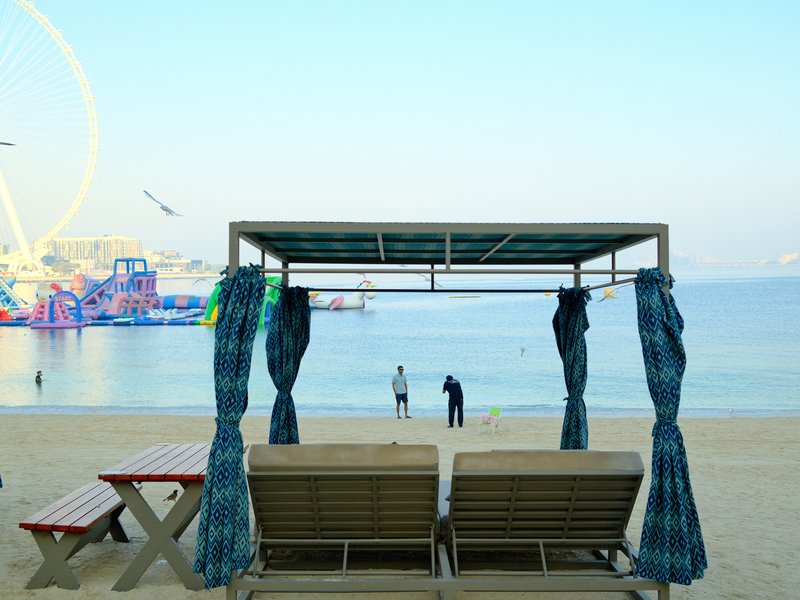 Two beach chairs under a canopy with a view of the sea and distant amusement rides.