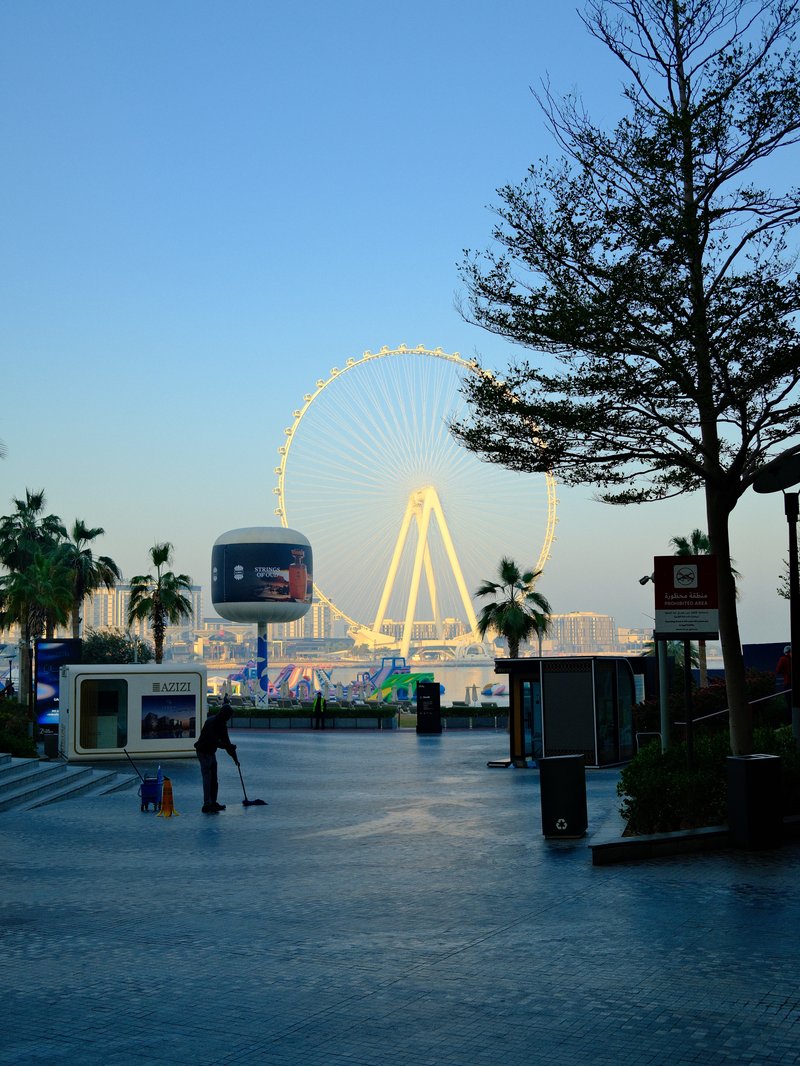 A quiet waterfront scene featuring a large Ferris wheel illuminated against a blue sky.