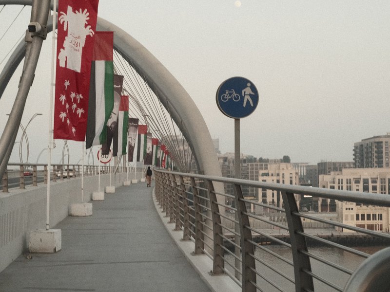 A pedestrian bridge with flags and signage, leading into an urban area at dusk.