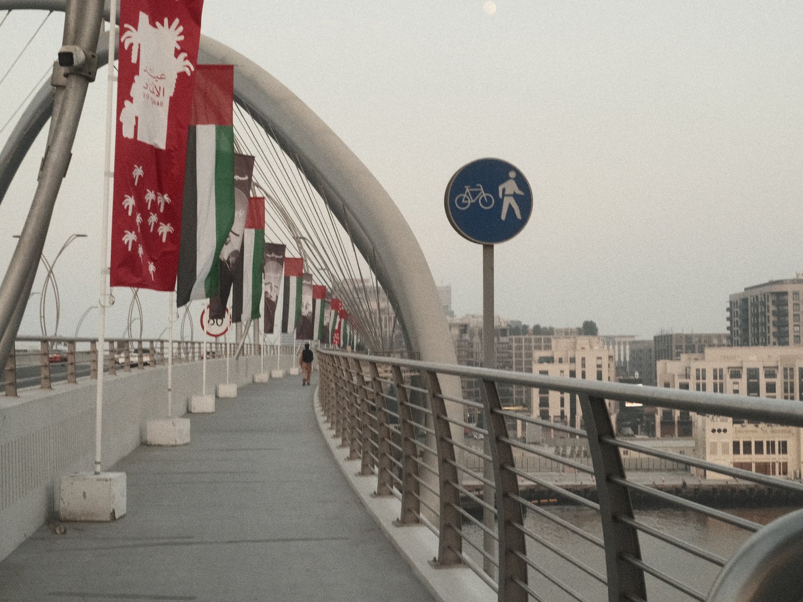 A pedestrian bridge with flags and signage, leading into a cityscape at dusk.