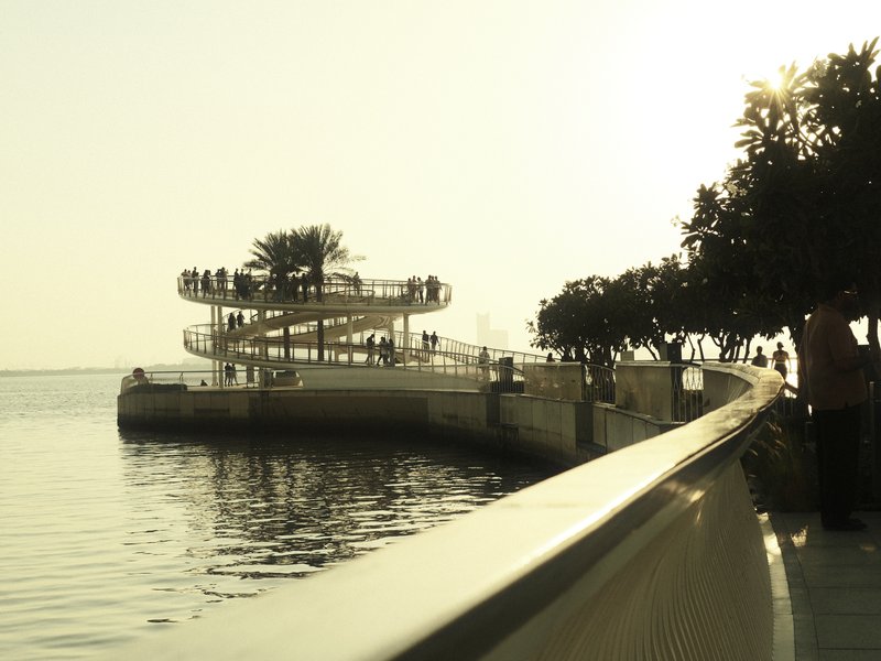 A winding walkway along a waterfront with a spiral structure and silhouetted trees at sunset.