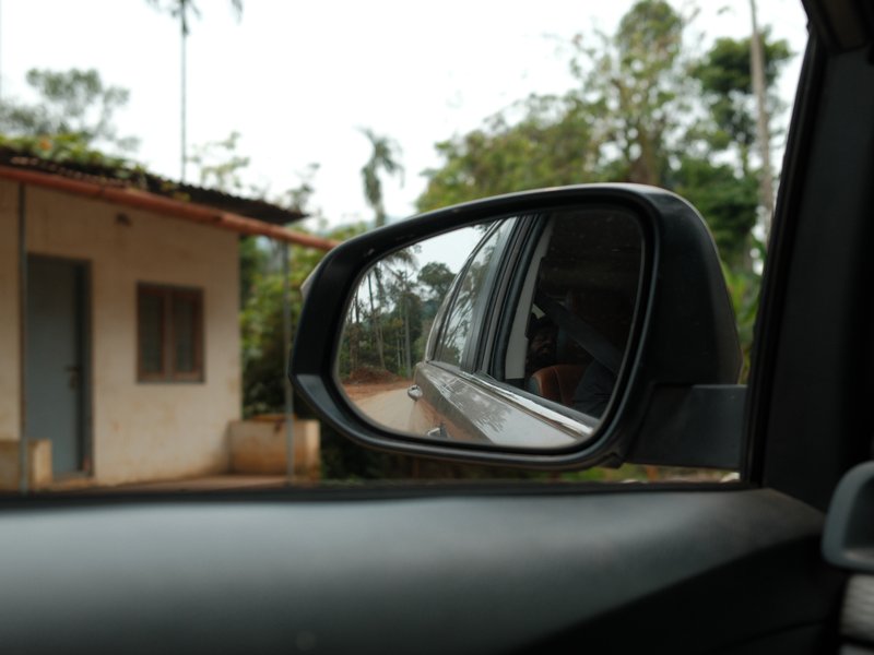 Side mirror of a car reflecting a rural scene with a house and trees in the background.