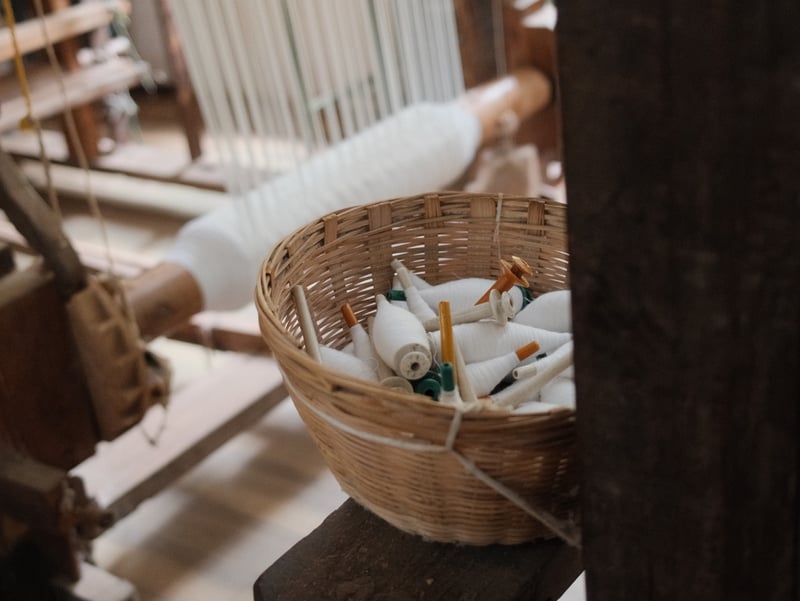 A woven basket filled with spools of thread in a weaving workshop, with looms in the background.