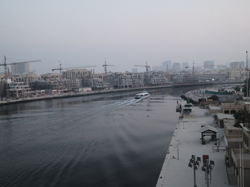 A boat gliding through a calm river with urban buildings and cranes in the background under a hazy sky.