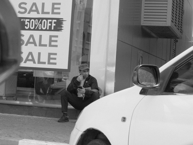 A man sitting on the sidewalk near a store with a large sale sign, partially obscured by a car.