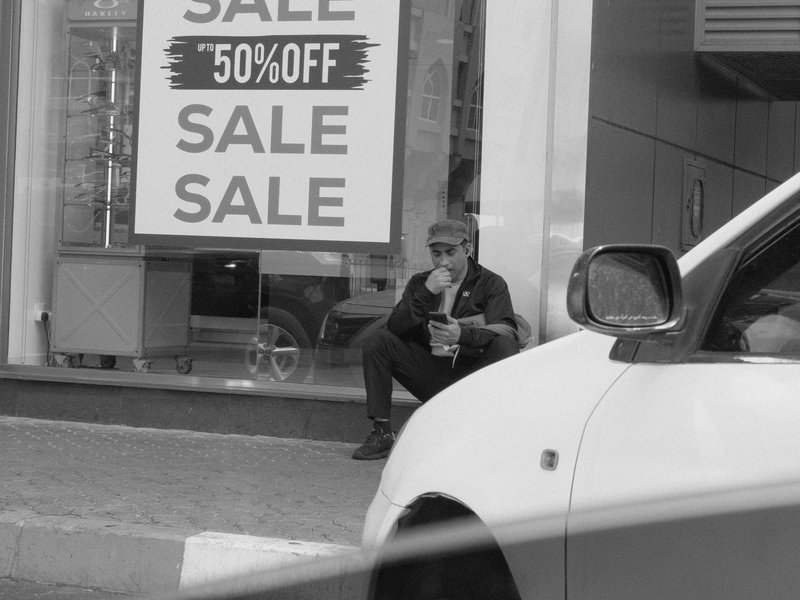 A man sitting on the sidewalk near a store with a sale sign, sipping a drink.