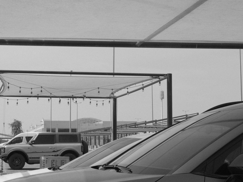 A black and white photo of parked vehicles under a canopy with string lights in the background.