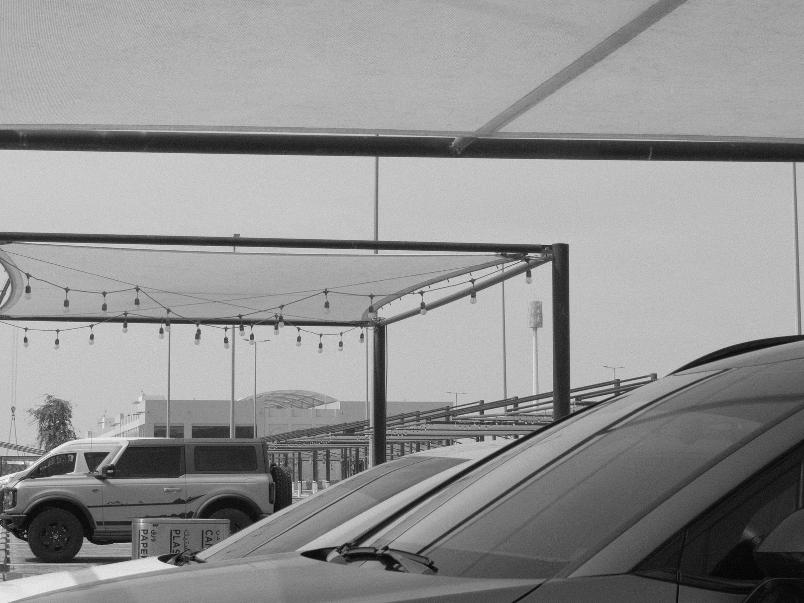 A black and white photo of parked vehicles under a canopy with string lights in the background.
