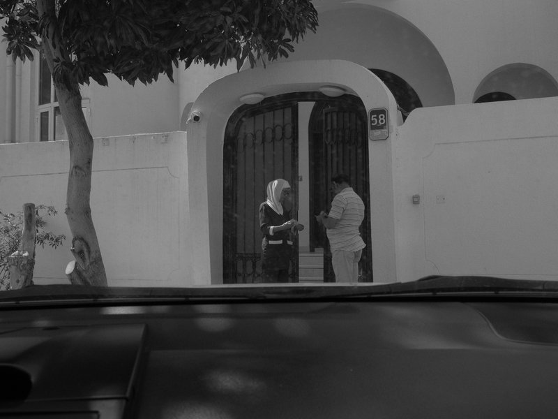 Two people conversing outside a white wall with a gate, viewed from inside a car.