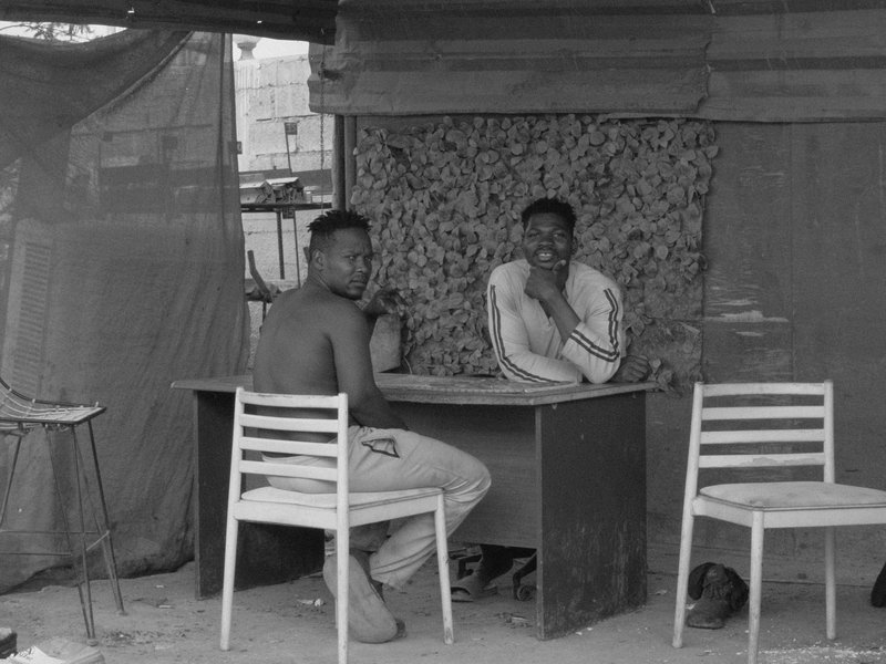 Two men sitting at a table in a makeshift outdoor setting, with a textured background.
