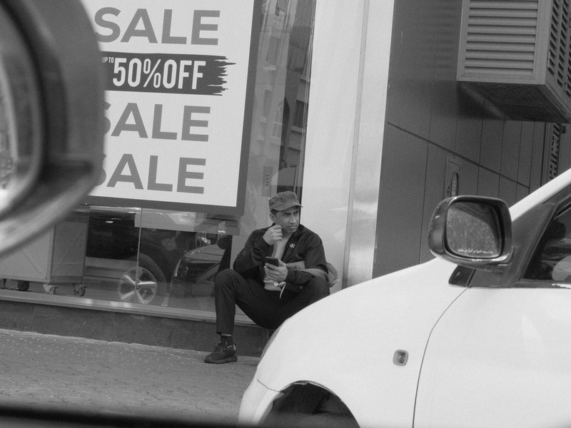 A man sitting on the sidewalk next to a large sale advertisement, partially obscured by a car.