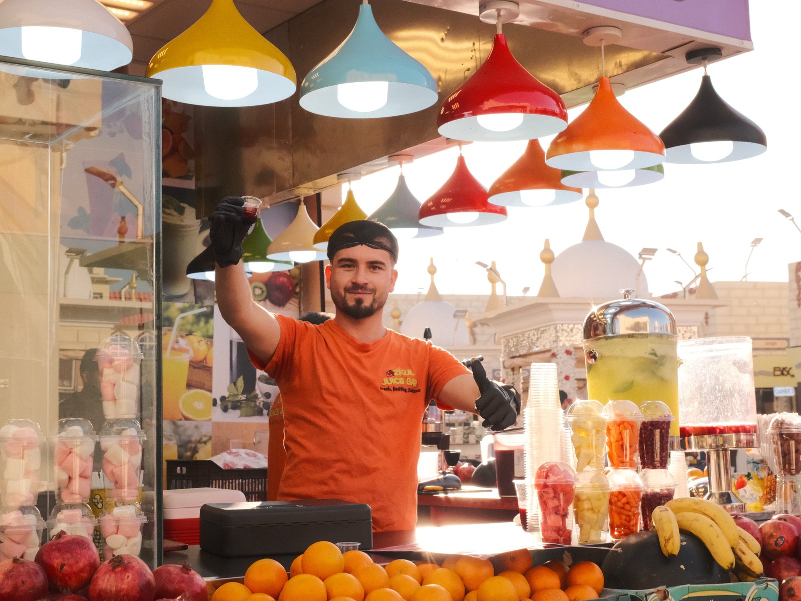 A vendor in an orange shirt stands behind a colorful fruit stall with various fruits and drinks.