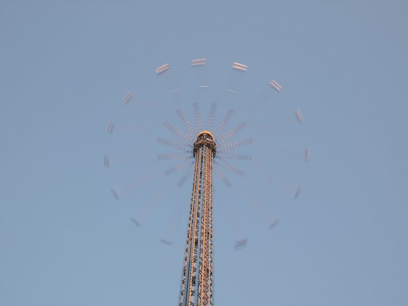 A tall amusement park ride against a clear blue sky, with colorful seats circling at the top.