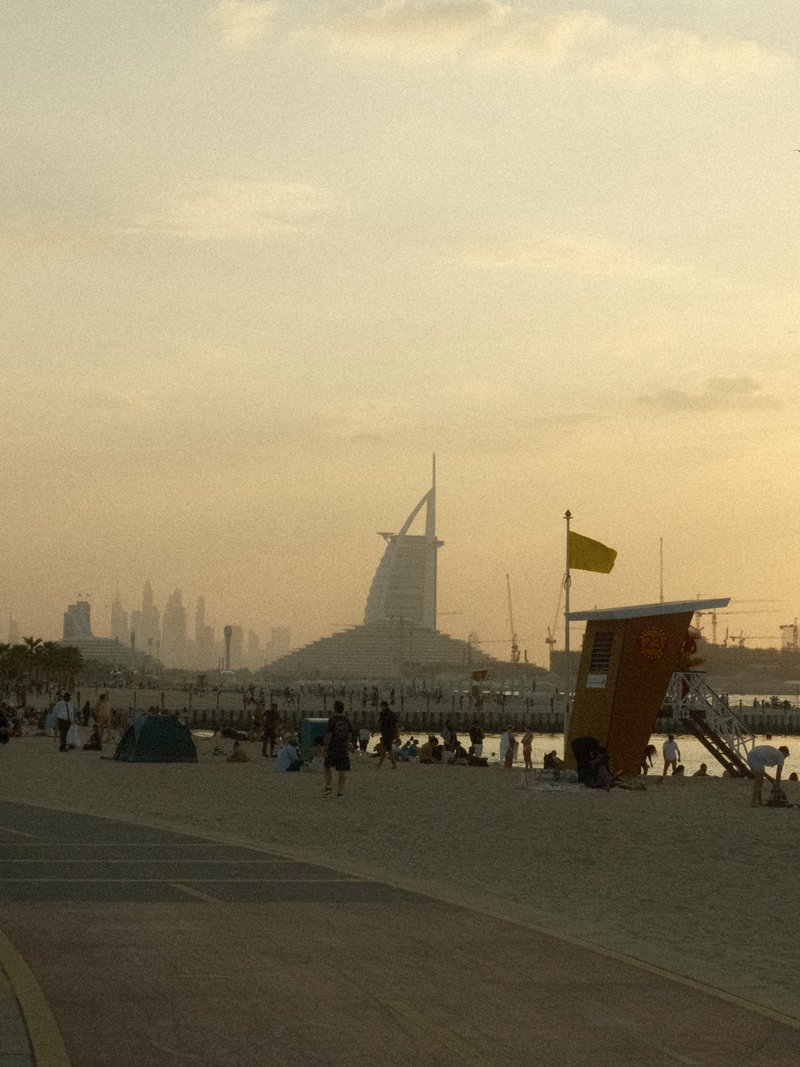 Silhouette of a beach scene at sunset with people, lifeguard tower, and distant city skyline.