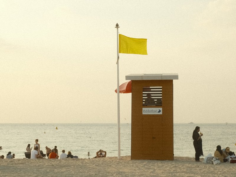 A lifeguard station on a beach with a yellow flag, people relaxing on the sand, and a calm sea in the background.