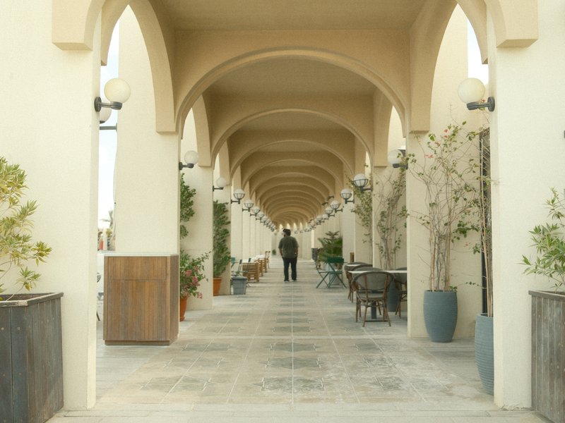 A person walking through a long corridor with arches and potted plants.