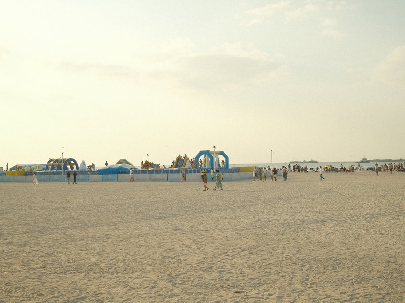 A beach scene with people walking on sand, inflatable attractions in the background under a hazy sky.