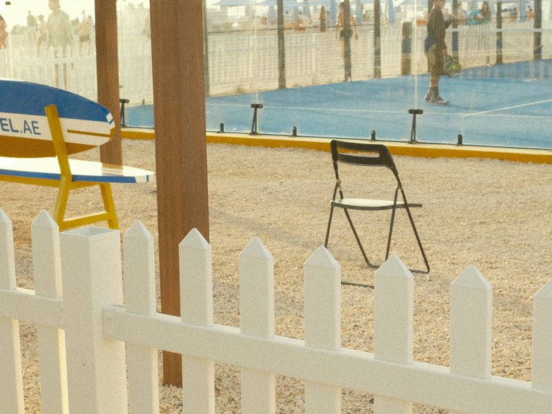 A vacant chair in front of a tennis court, with a white picket fence and a blue sky in the background.