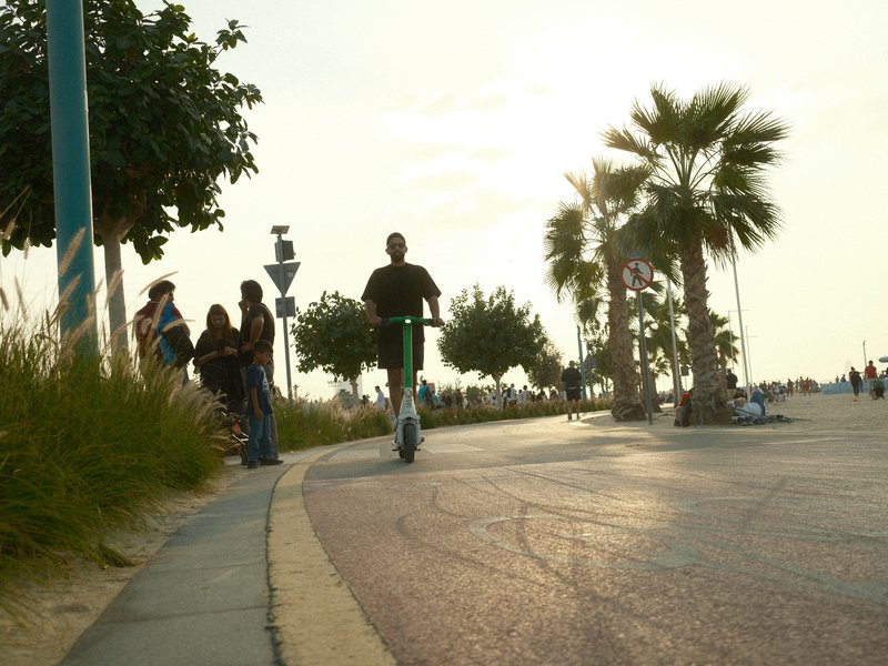 A person riding a scooter along a palm-lined path with people in the background during sunset.