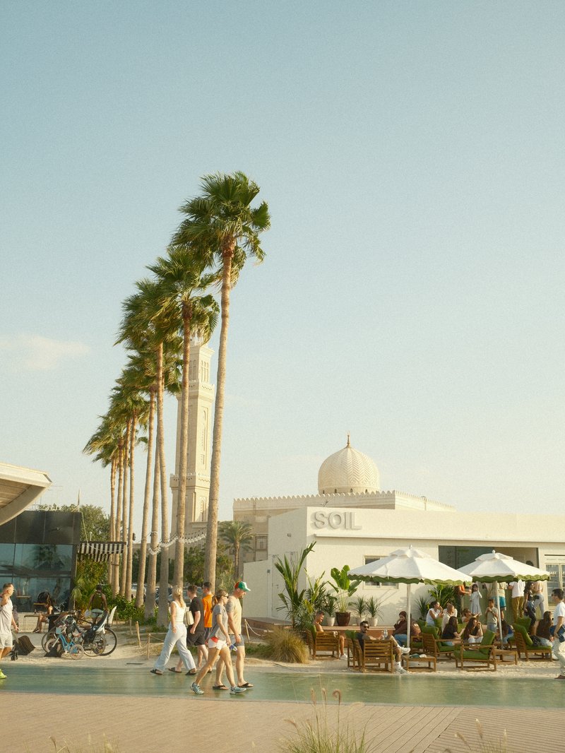 A sunny outdoor scene with palm trees, people walking, and a white building with a dome.