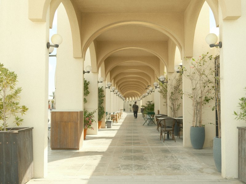 A person walking through a sunlit corridor with arches and potted plants.