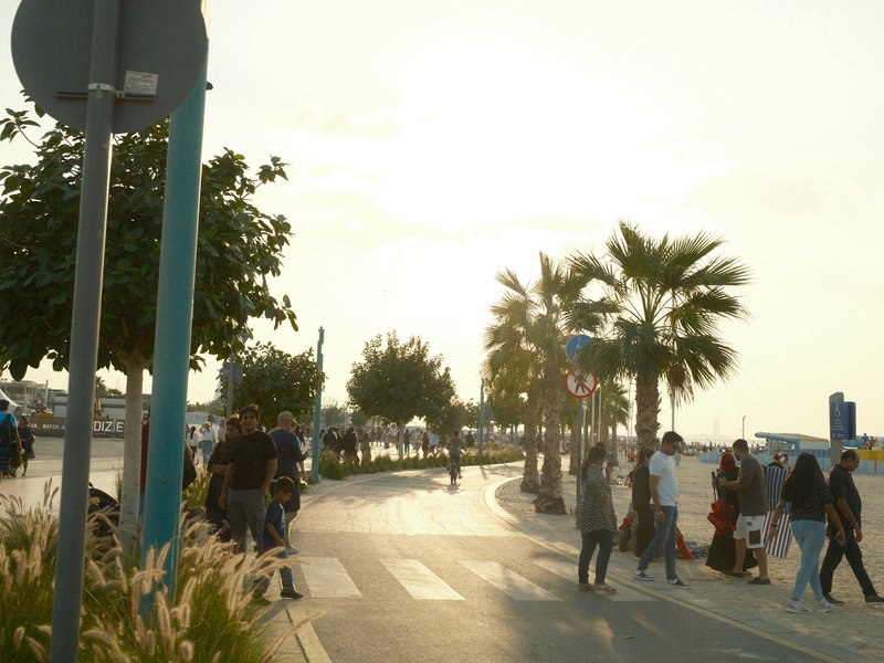 A lively promenade with people walking, palm trees, and a bright sun setting in the background.