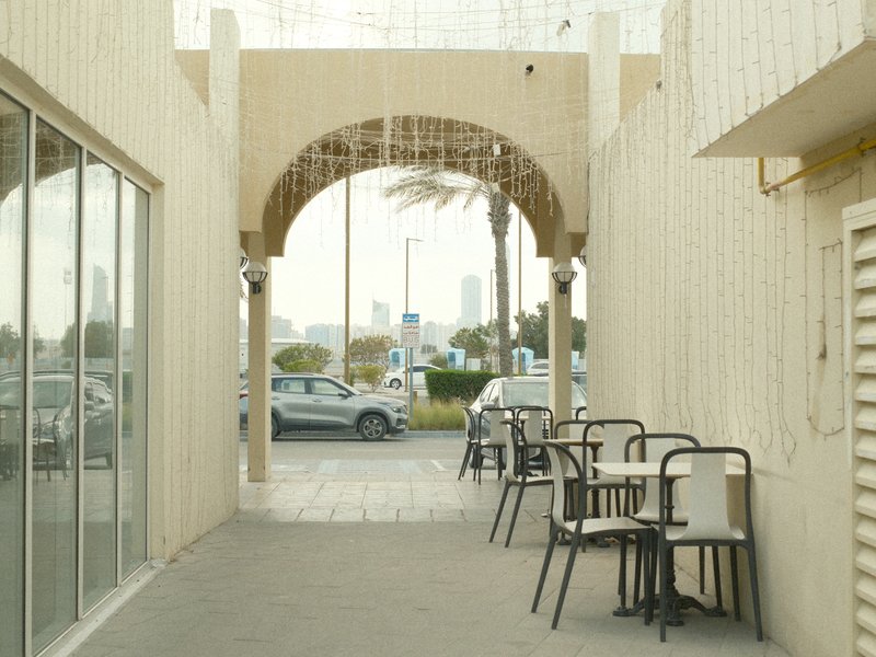 A narrow walkway with empty tables and chairs, leading to a street view with palm trees and buildings in the distance.