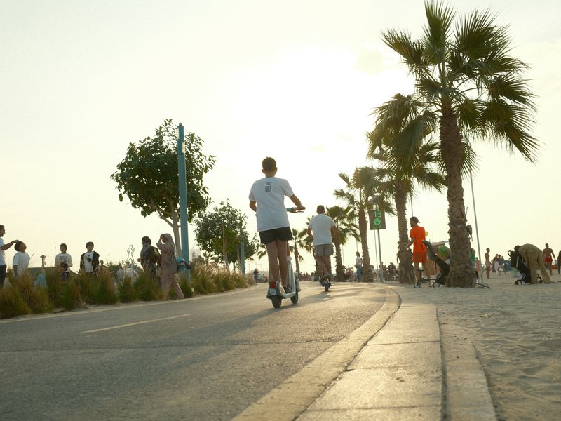 A person riding a scooter along a palm-lined path during sunset with people in the background.