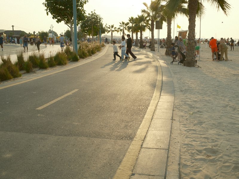 A sunlit promenade with people walking, palm trees lining the path, and a sandy beach in the background.