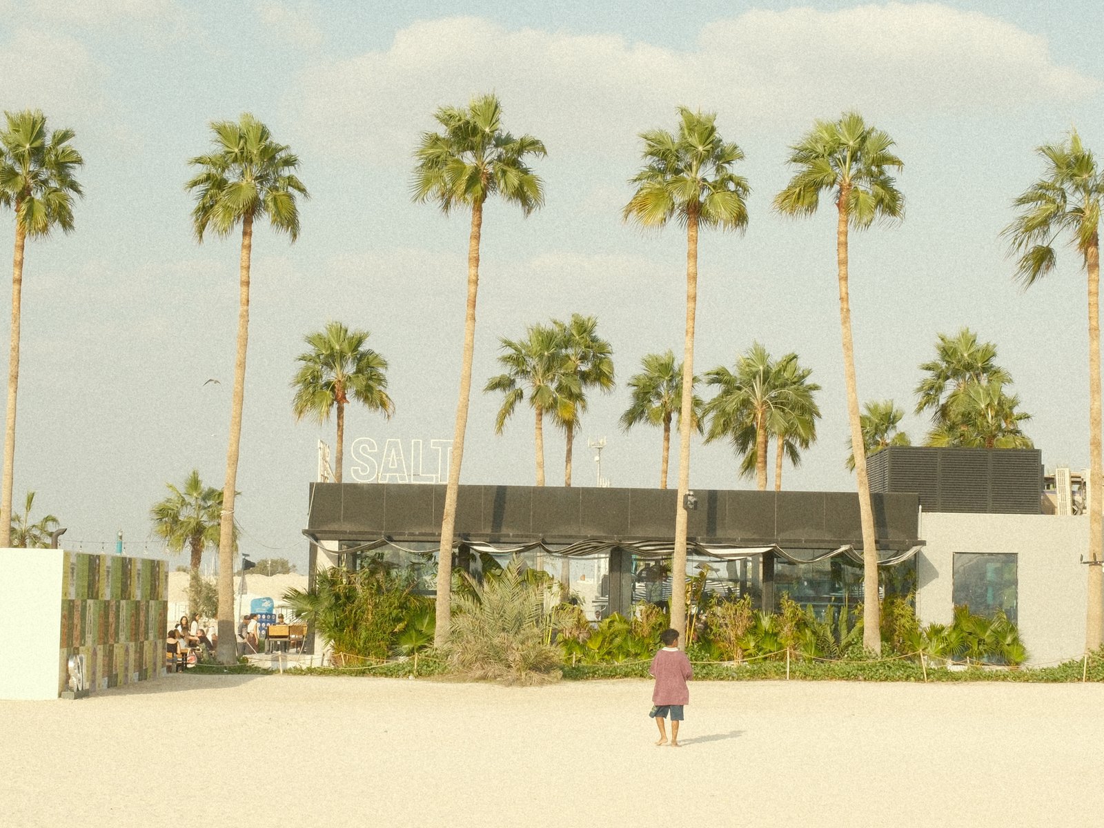 A person walking on a sandy beach with palm trees and a modern building in the background under a bright sky.