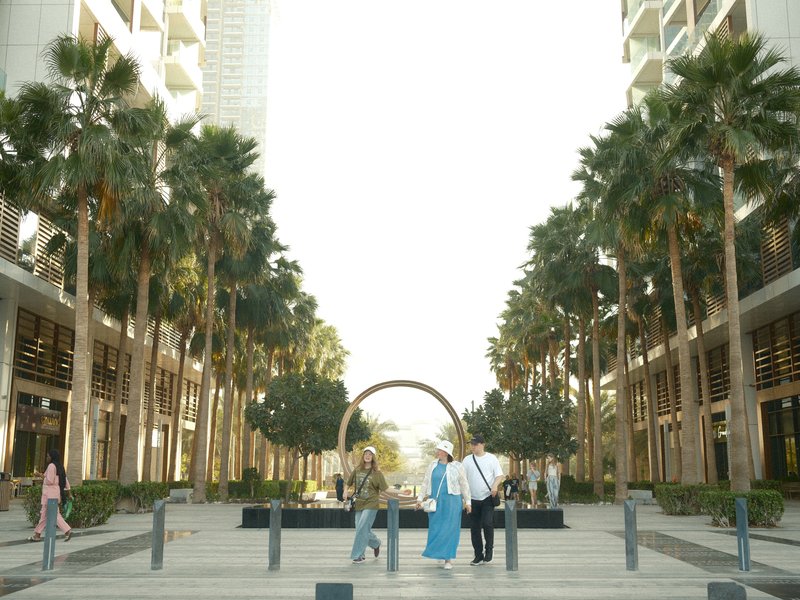 A bustling urban plaza lined with palm trees and modern buildings, featuring people walking and socializing.