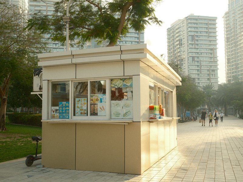A small kiosk with large windows in an urban park setting, surrounded by tall buildings and people walking.
