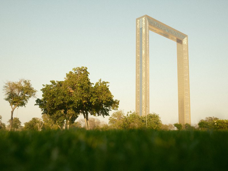 A tall, modern structure resembling a frame stands against a clear sky, surrounded by trees and grass.
