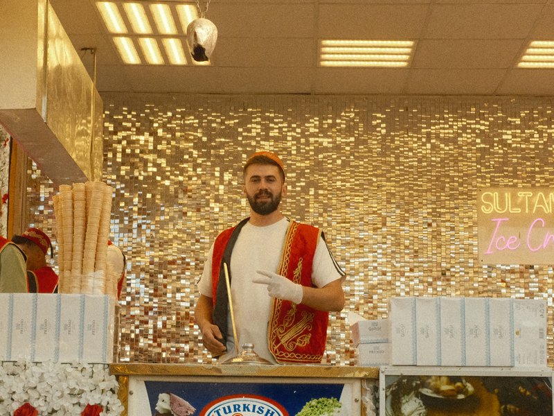 A vendor in a bright shop with a sparkling background, wearing a red vest and making a peace sign.