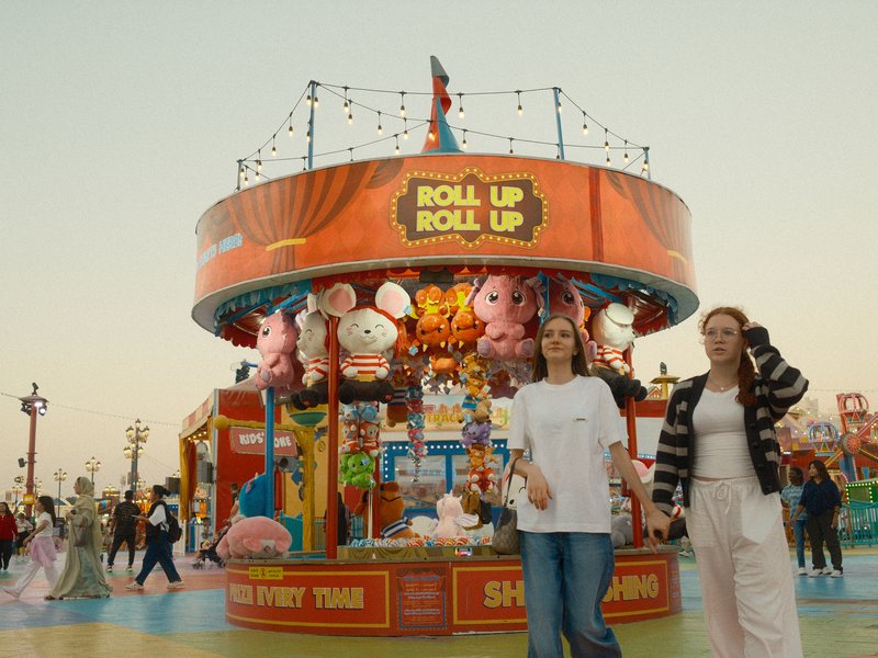 Two young women stand in front of a colorful carnival game booth at sunset.