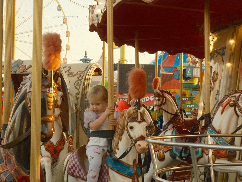 A young child sitting on a carousel horse at a carnival, with colorful lights and decorations in the background.